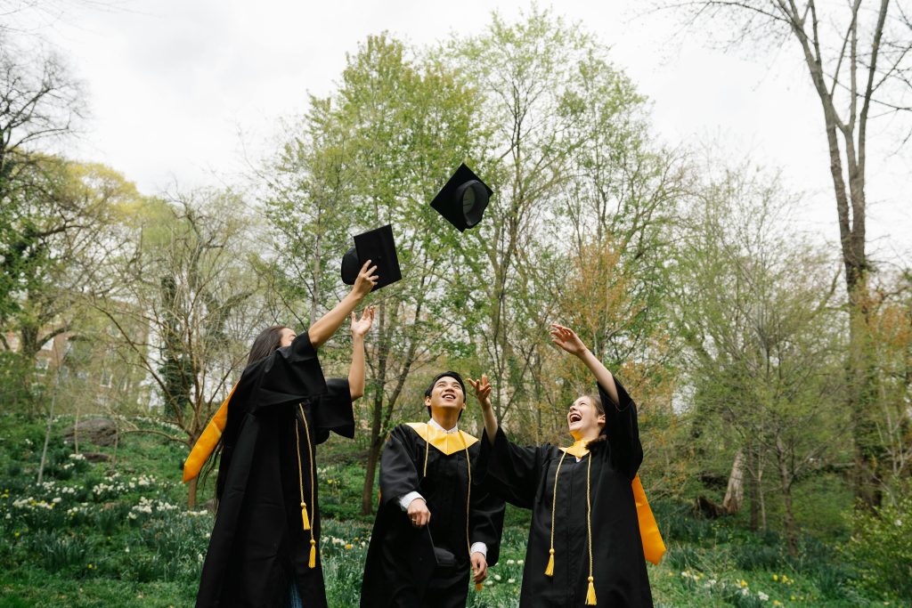 Three graduates celebrating and throwing caps in the air in a lush green park.