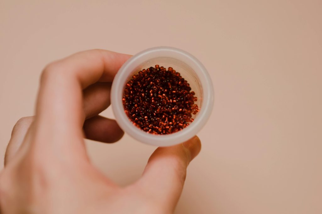 Close-up of a hand holding a round container filled with small red beads, perfect for crafts.