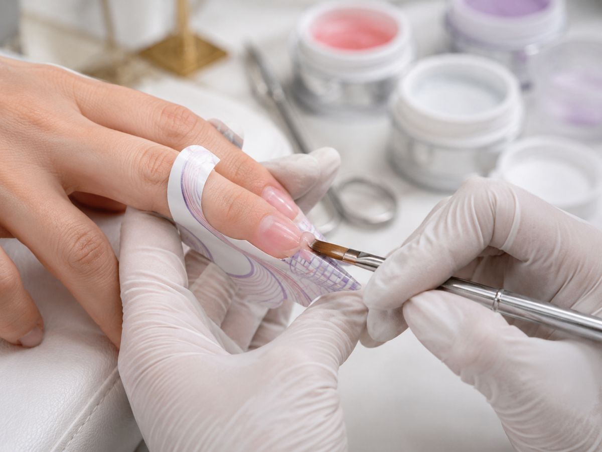 Kozmetičarka s bijelim rukavicama nanosi gel kistom na produžetak deformiranog nokta pomoću papirnate šablone, dok je ruka klijentice oslonjena na jastučić