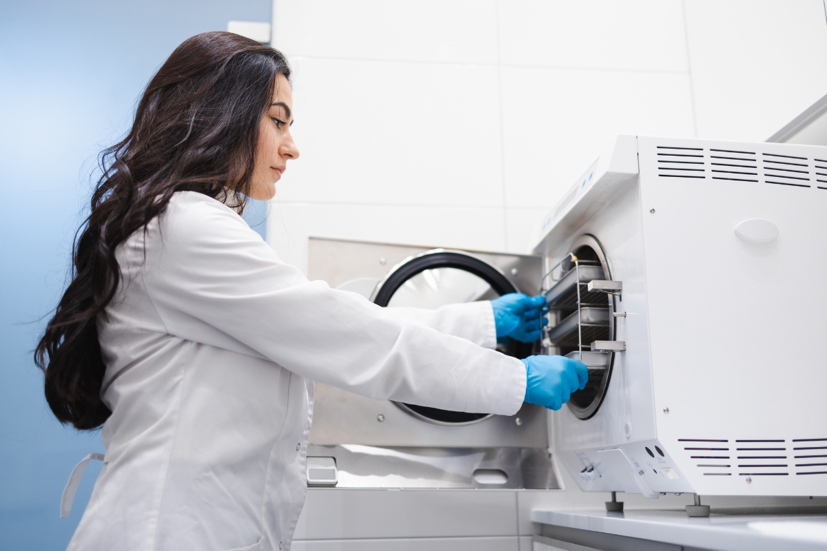 Žena u laboratorijskom okruženju stavlja opremu u sterilizator (autoklav), noseći zaštitne rukavice.