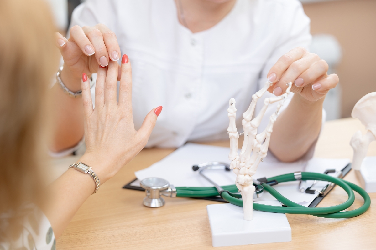 Zdravstveni djelatnik pregledava pokretljivost i funkciju prstiju šake pacijentice, uz anatomski model kostiju šake na stolu tijekom pregleda mišićno-koštanog sustava.