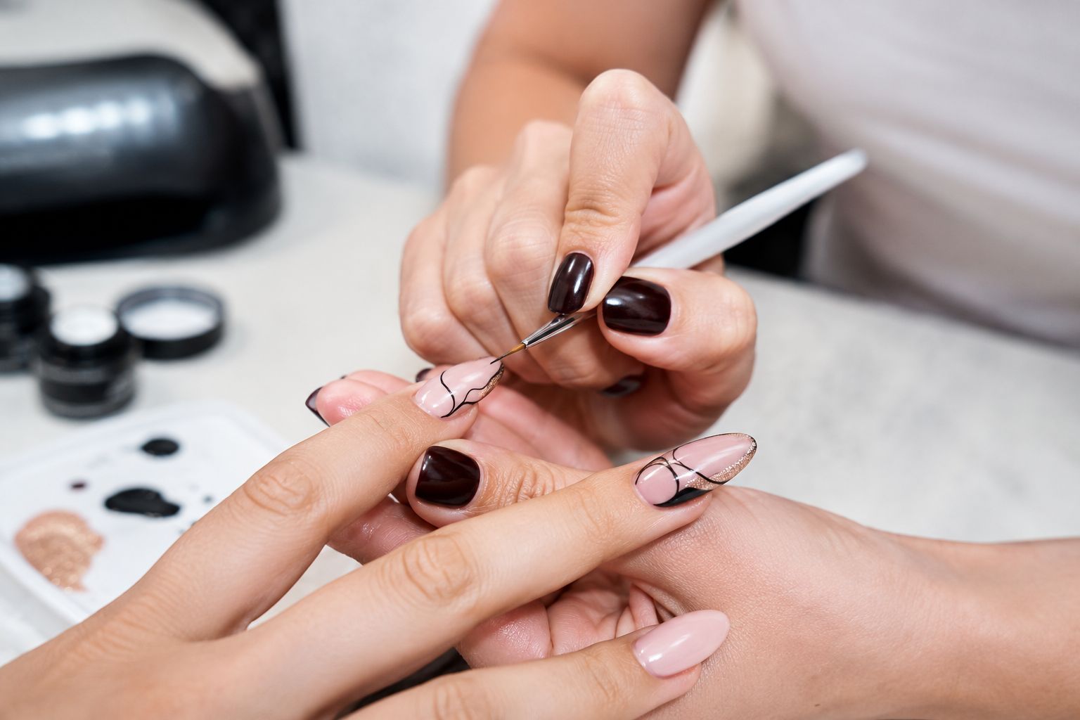 Kozmetičarka u salonu tankim kistom izrađuje nail art na bademasto oblikovanim noktima klijentice, s preciznim linijama i sjajnim detaljima u nježnim nude tonovima