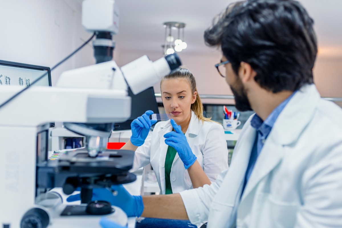 Dvoje laboratorijskih stručnjaka u bijelim kutama rade u suvremeno opremljenom laboratoriju. Žena s plavim zaštitnim rukavicama pažljivo pregledava stakalce za mikroskop, dok njezin kolega upravlja mikroskopom. U prvom planu nalazi se laboratorijska oprema, a prizor prikazuje znanstveno istraživanje, analizu uzoraka i timski rad u laboratorijskom okruženju.