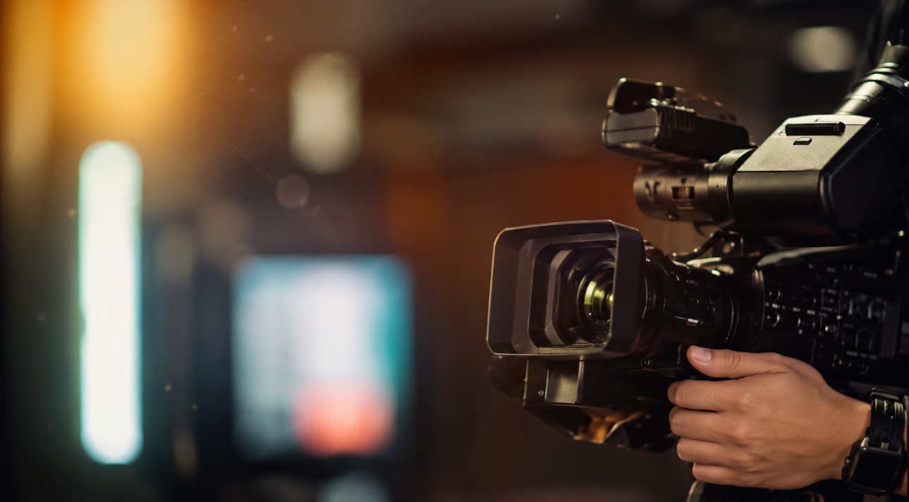 Krupni kadar profesionalne video kamere u rukama snimatelja, uz toplo studijsko osvjetljenje i zamućenu pozadinu
